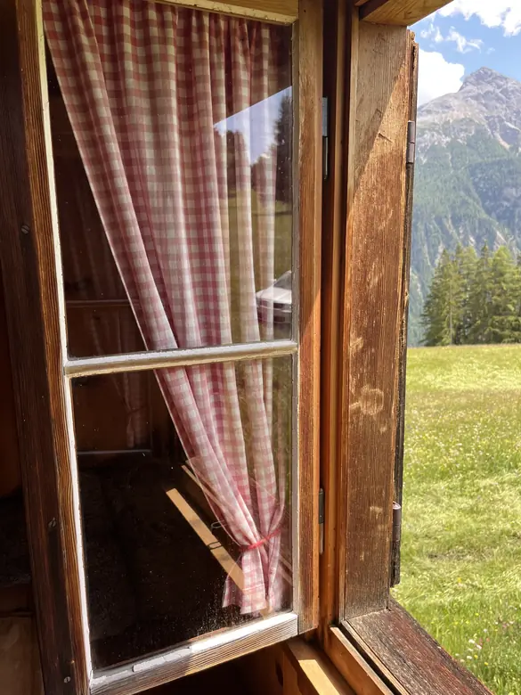 Offenes Fenster mit rot karierten Vorhängen, draussen Wiese, Fichten und Berge