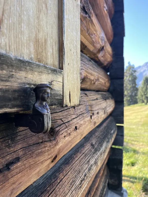 Alter Halter aus Eisen in Form einer Figur an der verwitterten Balkenwand, dahinter Wiese und Fichten