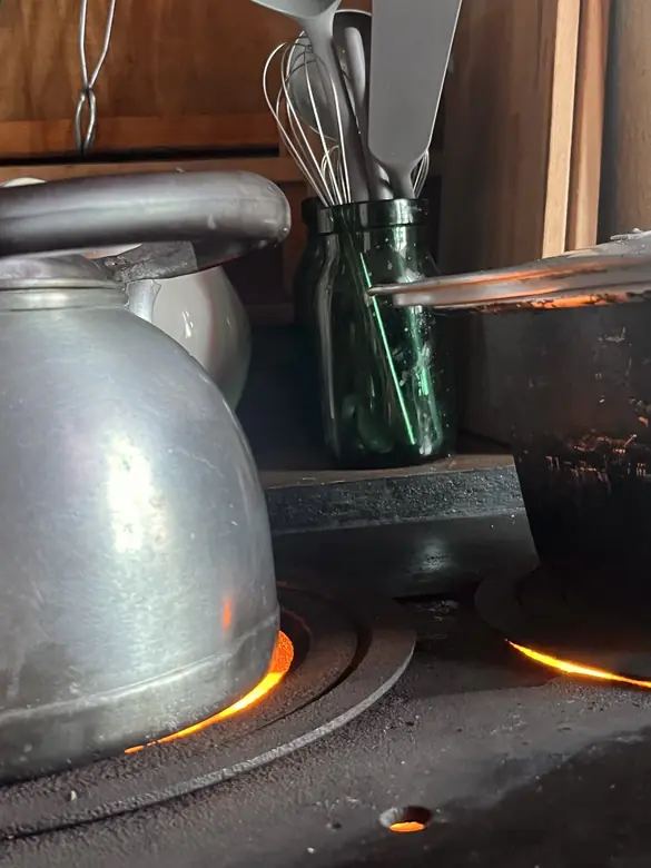 Wasserkessel auf dem Holzherd, die Glut leuchtet durch die Ringe, dahinter Schwingbesen in einem grünen Glas