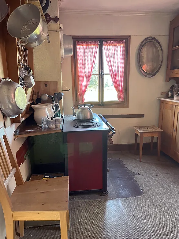 Küche mit Holzherd, Pfannen an der Wand, Wasserkessel und Fenster mit rot karierten Vorhängen