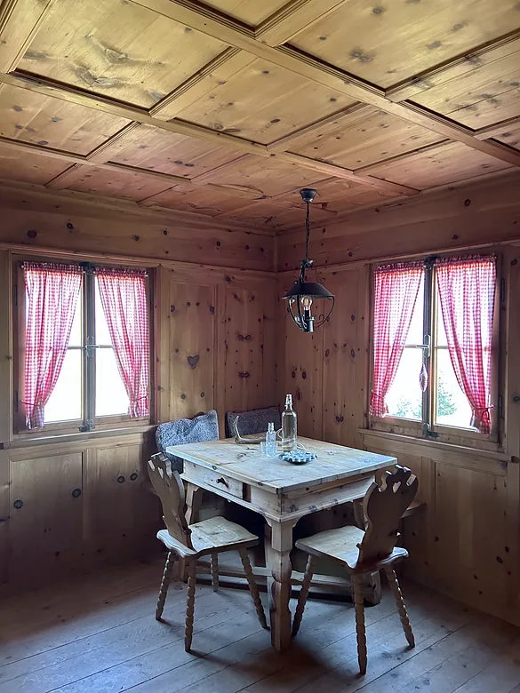 Arvenstube mit Kassettendecke, Holztisch, Stühlen und zwei Fenstern mit rot karierten Vorhängen